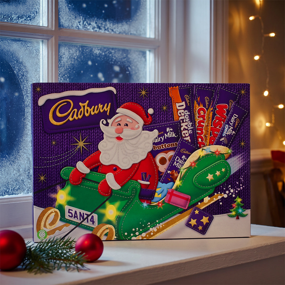 Cadbury advent calendar with a Santa design on a windowsill with a snowy window, baubles, and fairy lights