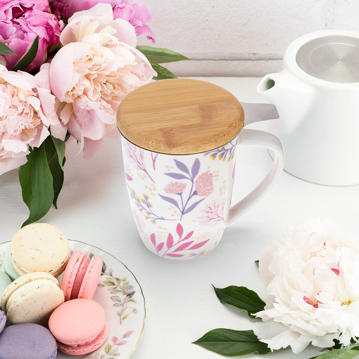 Floral mug with wooden lid, teapot, and macarons on a white surface with flowers.