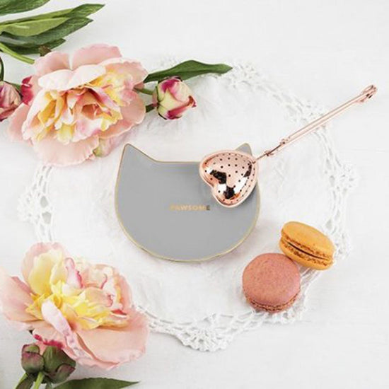 Cat-shaped tea infuser caddy with rose gold accents on a white lace doily with flowers and macarons and a heart shaped tea infuser.