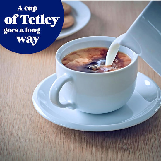 Light blue teacup filled with Tetley extra strong tea on a table with cream being poured in from a light blue creamer. A light blue plate of cookies is in the far-right corner covered with a blue circle call out for the product.