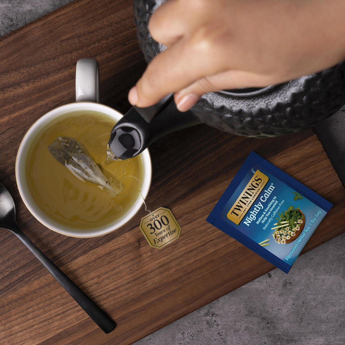Person pouring tea from a black teapot into a white cup on a wooden surface, with a Twinings tea bag and teabagging tool visible.
