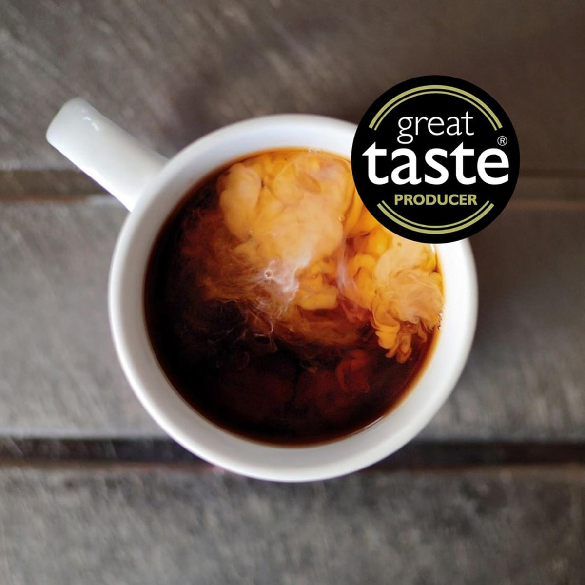 White teacup filled with hot tea and cream on a wooden surface, featuring the 'Great Taste' producer logo.