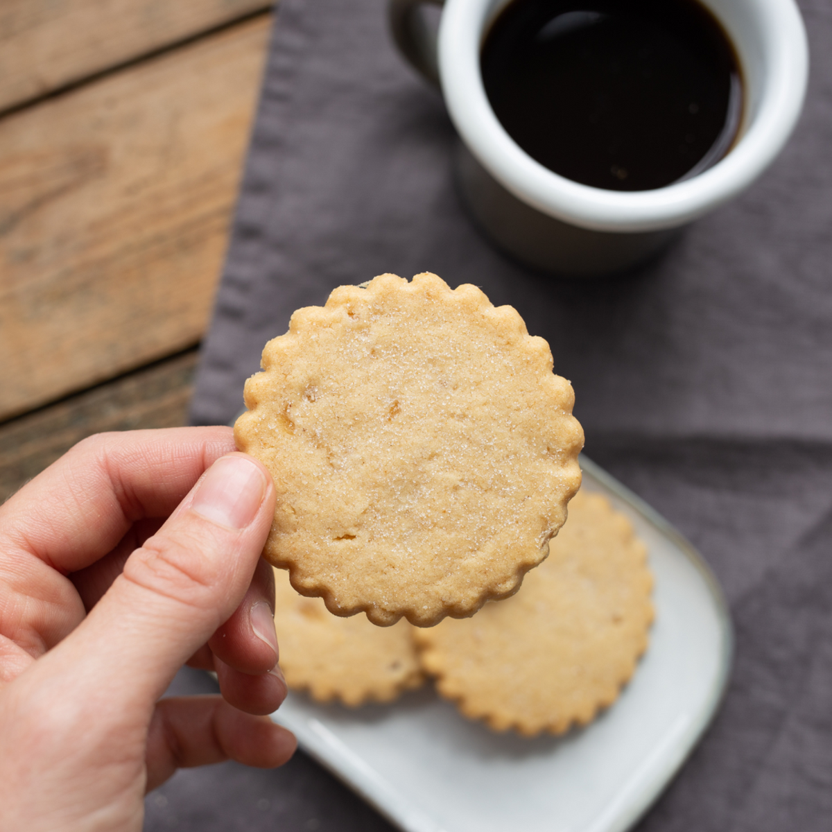 Hand holding a round Walker's shortbread cookie with a cup of coffee in the background on a wooden surface.