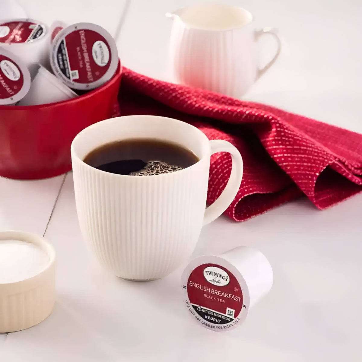 White mug with coffee, Twinings tea bags, and a red container on a white surface.
