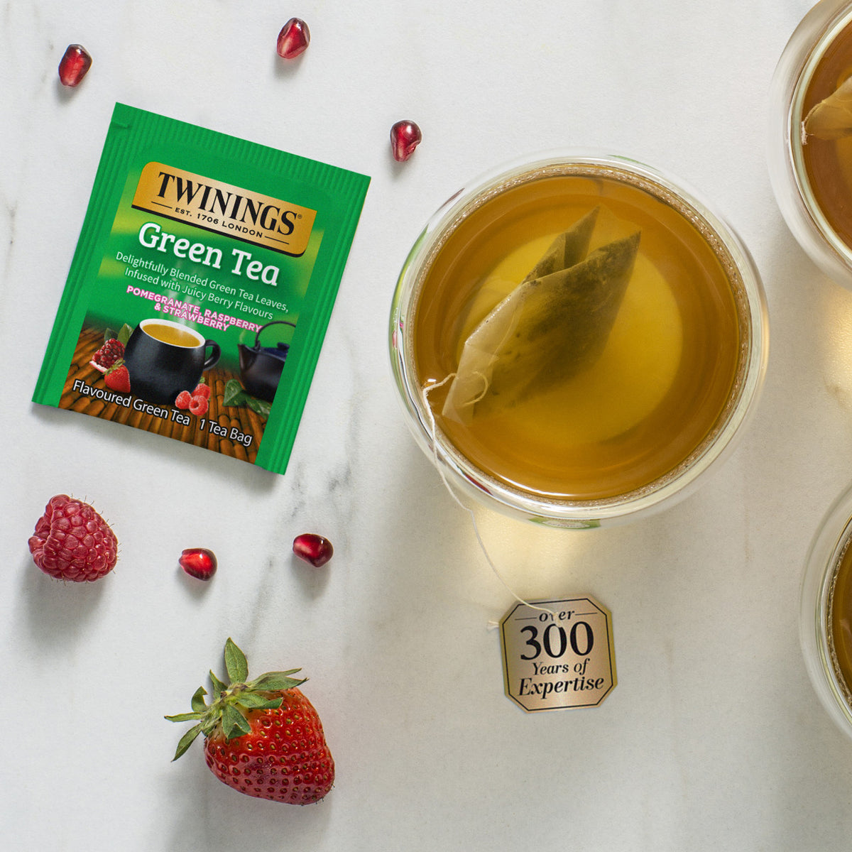 A Twinings green teabag package on a table with strawberries and cups of hot green tea with a teabag inside.