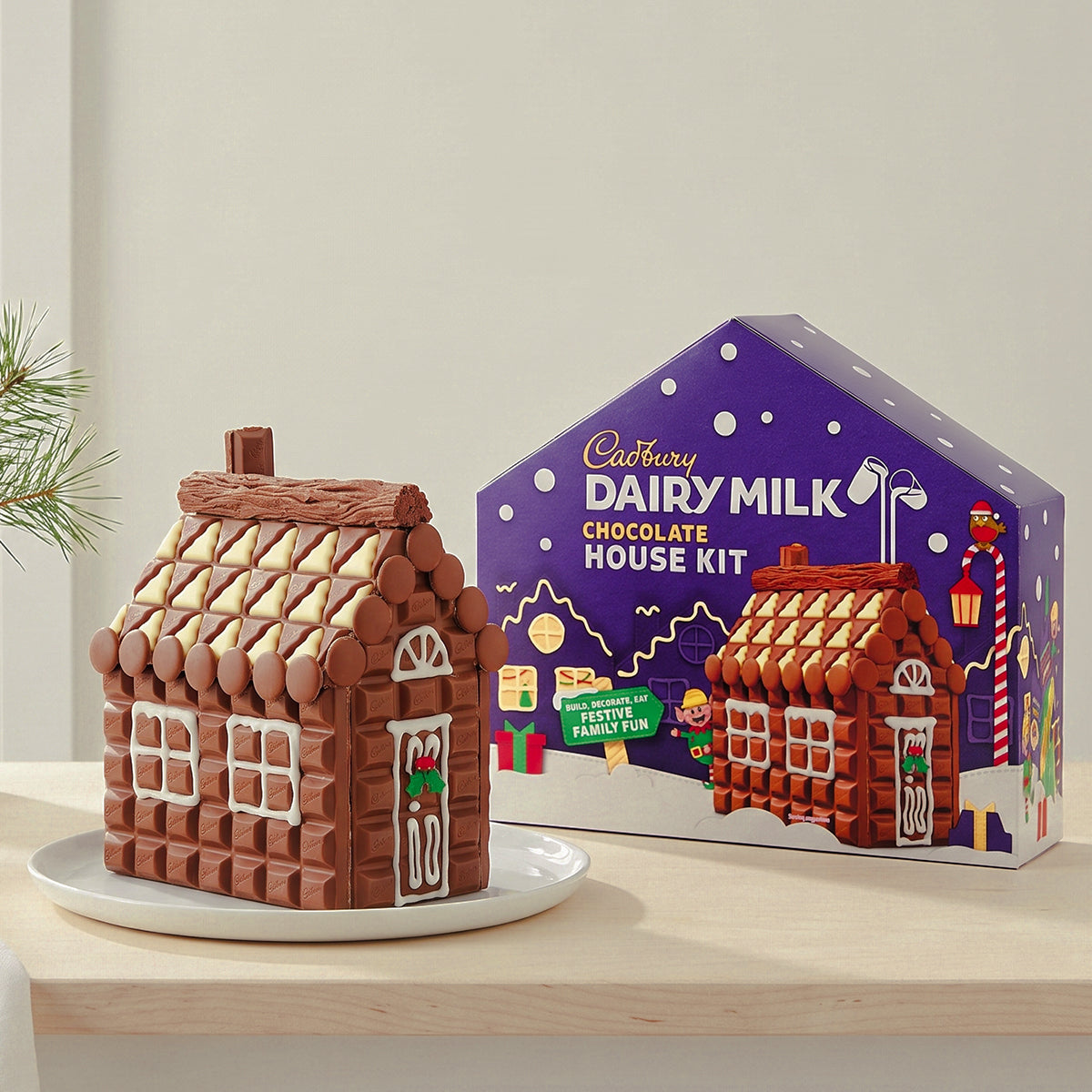 Finished chocolate house on a plate beside its Cadbury Dairy Milk House Kit box