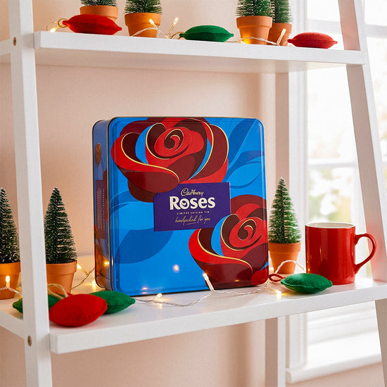 Cadbury Roses box on a white shelf with a red mug, small Christmas trees, and decorations