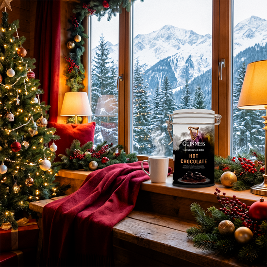 Guinness hot chocolate tin on a windowsill with a mug, a red throw, and a snowy mountain view