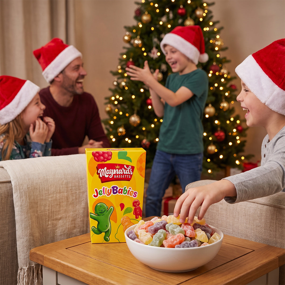Family wearing Christmas hats around a table with a bowl of jelly babies and a Maynards box, Christmas tree in the background.