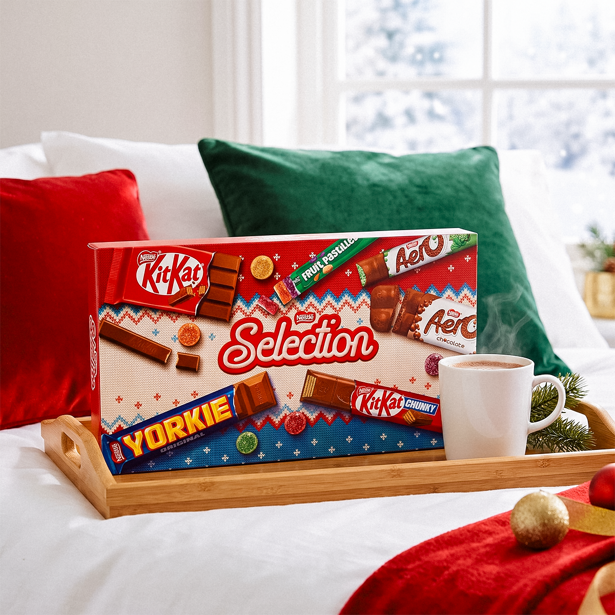 Nestlé Selection box on a tray with a mug, red and green pillows, and a snowy window behind