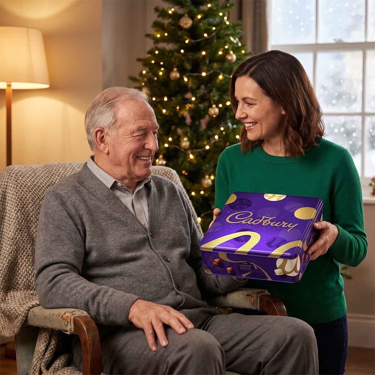Older man sitting in a chair as a woman hands him a Cadbury gift box, with a Christmas tree behind
