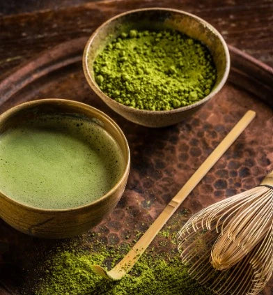 Matcha green tea in a bowl with a whisk and a bowl of matcha powder on a wooden surface.