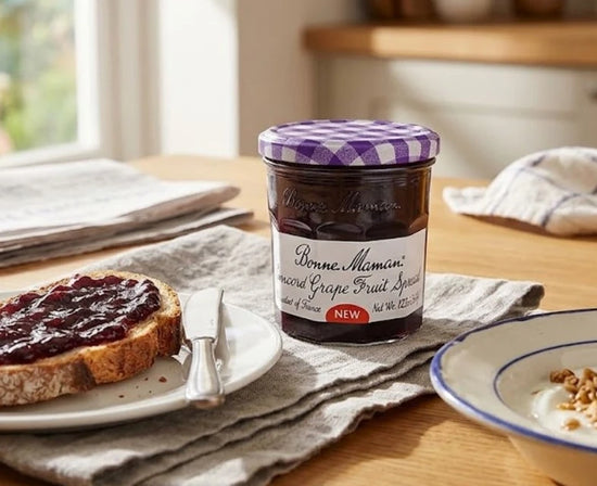 Jar of Bonne Maman grape fruit spread on a table with toast and a bowl.