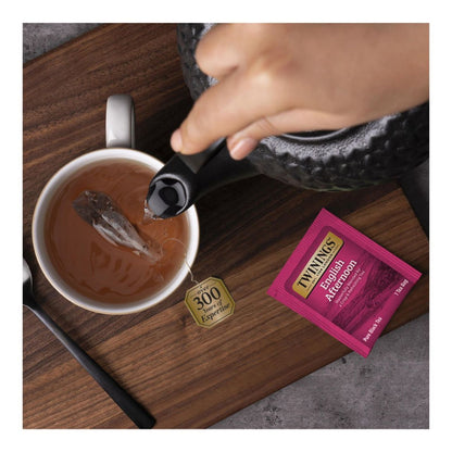 Tea being poured into a cup with a Twining's tea bag and teapot on a wooden surface.
