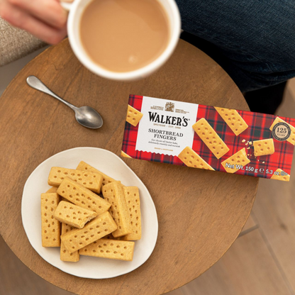 Image of a cup of tea with cream in the tea, a plate of Walker's Shortbread Fingers, and a package for Walker's Shortbread cookies along with a spoon on a wooden table.