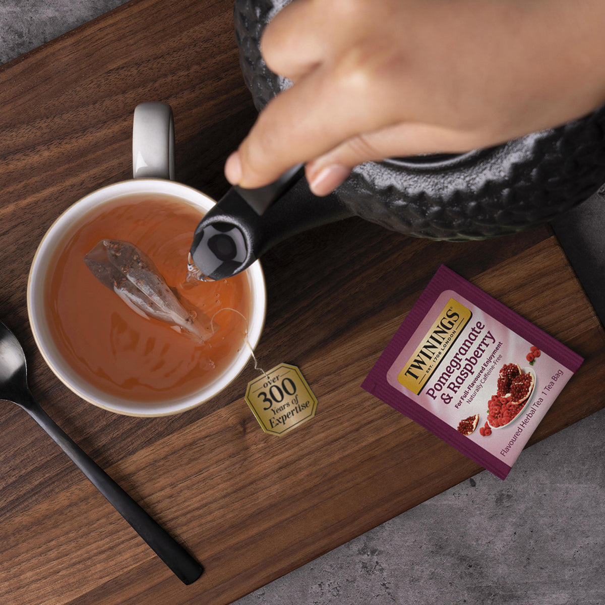 Tea being poured into a cup with a Twinings Pomegranate & Raspberry tea bag on a wooden surface.