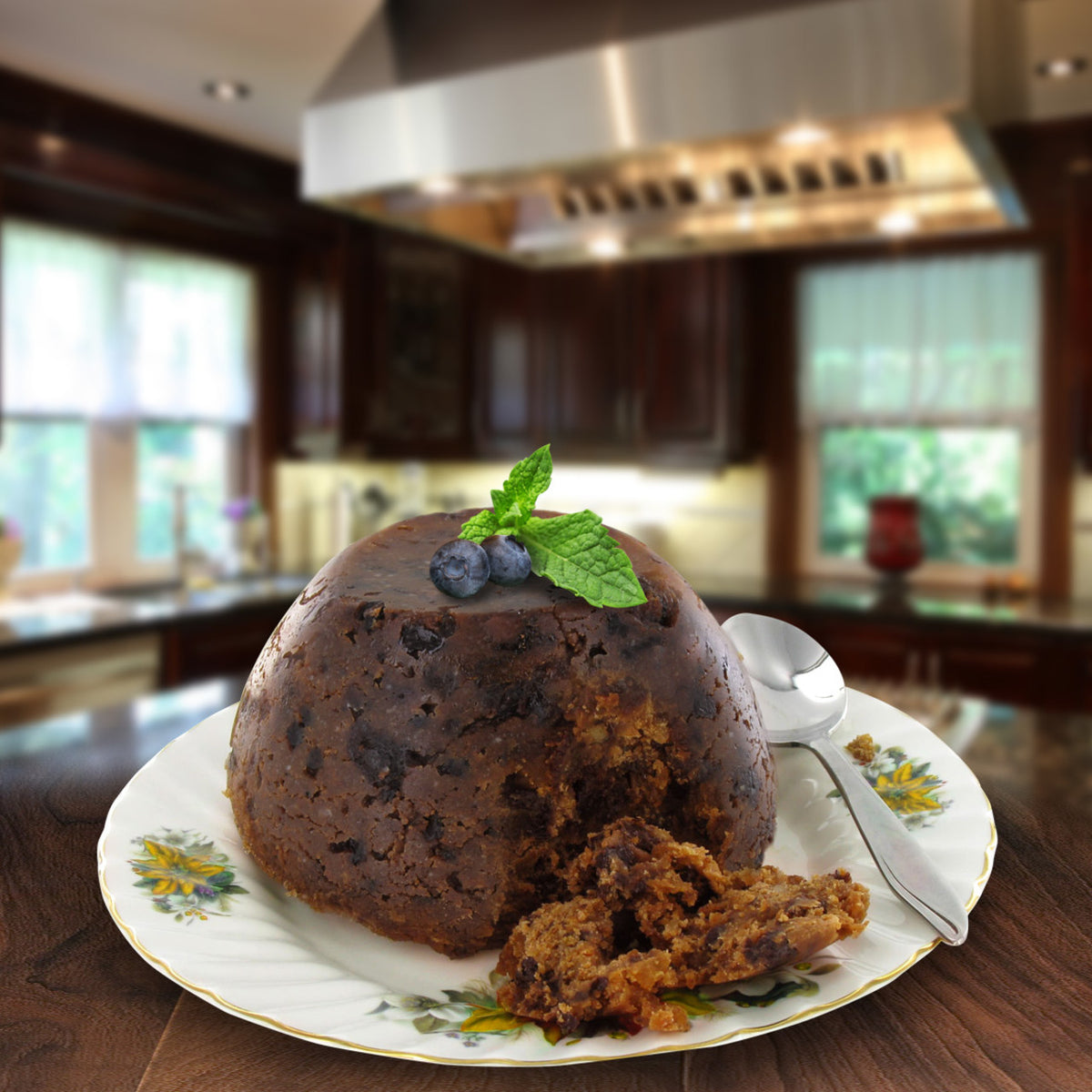 A Christmas Pudding baked desert on a floral plate with a sliver spoon.  The item super imposed on the backdrop of a modern looking kitchen featuring steel and dark wood.
