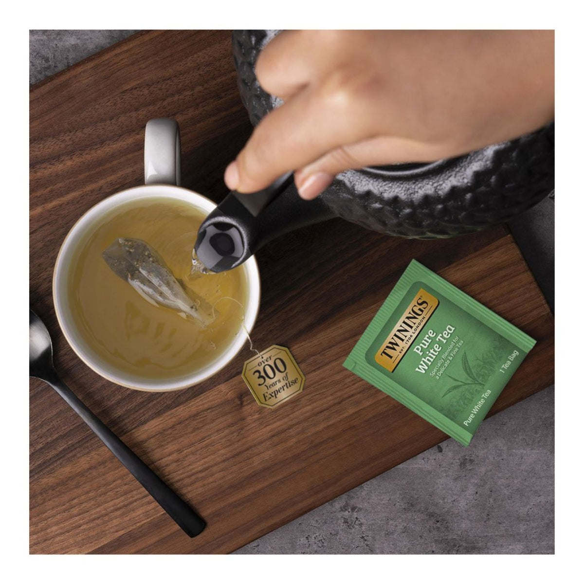 Twinings Pure White Tea being steeped in a white mug with a hand pouring water from kettle.  A greeen teabag package of Twinings Pure White Tea is on a wood tablet.
