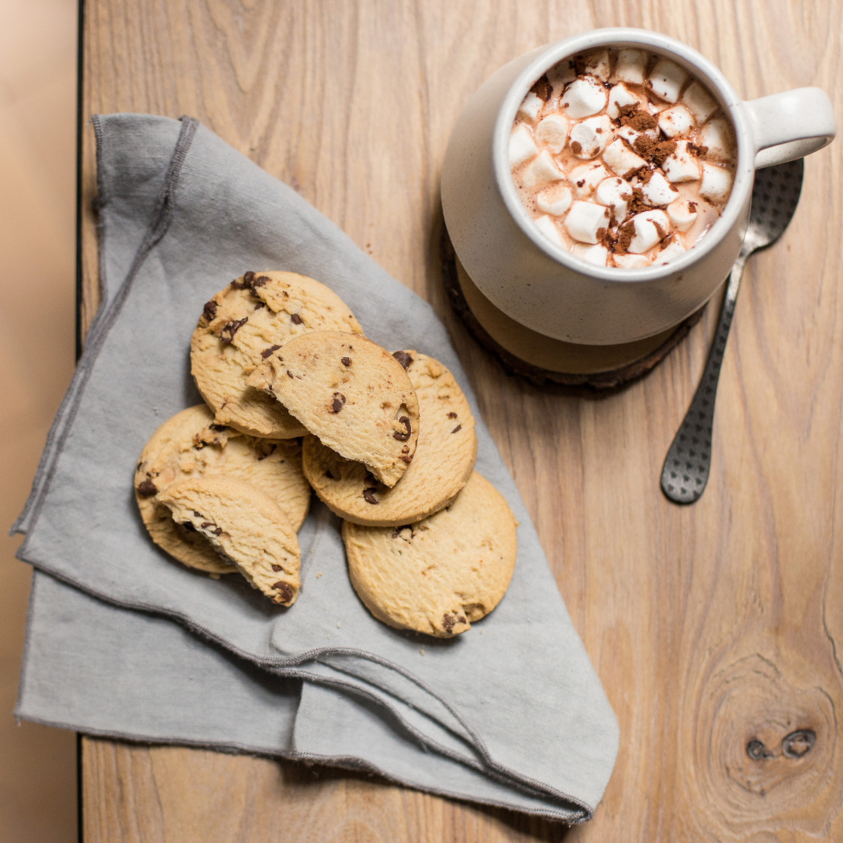 Walkers Shortbread Chocolate Chip Cookies on a gray napkin with a mug of hot chocolate topped with marshmallows on a wooden surface.