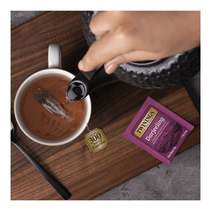 Person pouring tea into a cup with a Twining's tea bag and teapot on a wooden surface.