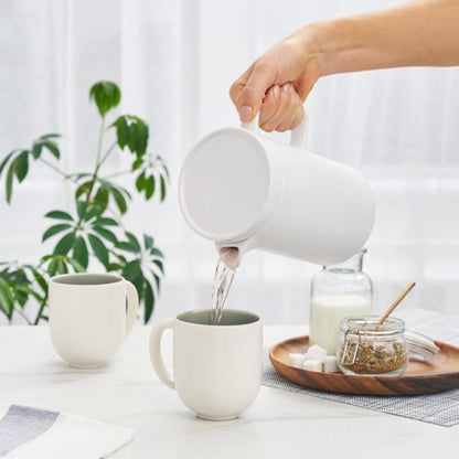 Person pouring liquid from a white pitcher into a white mug on a table with a plant and small items in the background.