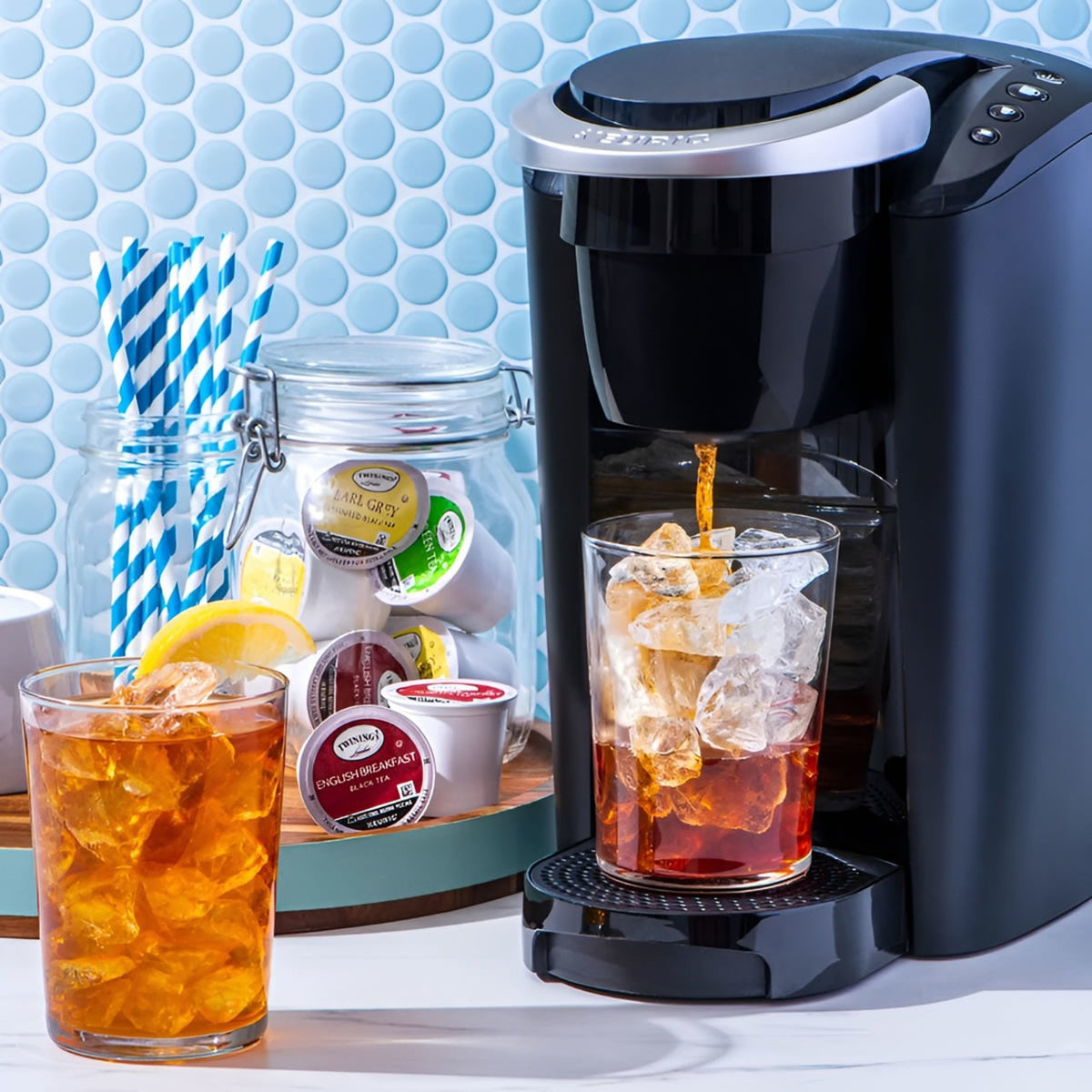 Black Keurig coffee maker pouring iced tea into a glass with lemon slices, against a light blue tiled wall.