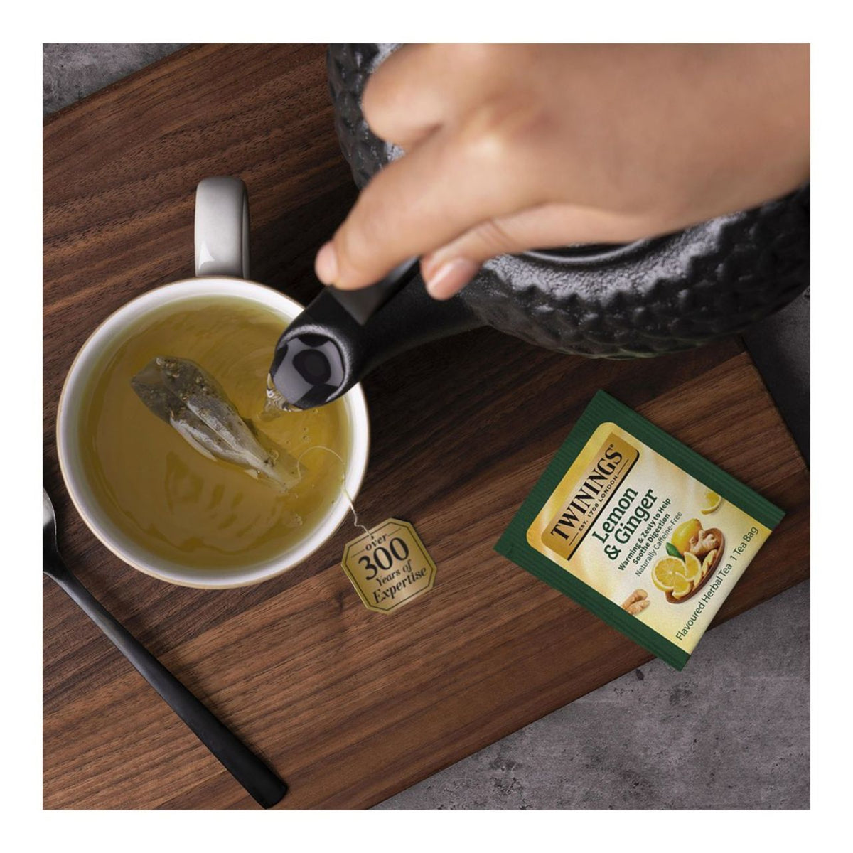 Person pouring tea into a cup with a Twining's Lemon & Ginger tea bag on a wooden surface.