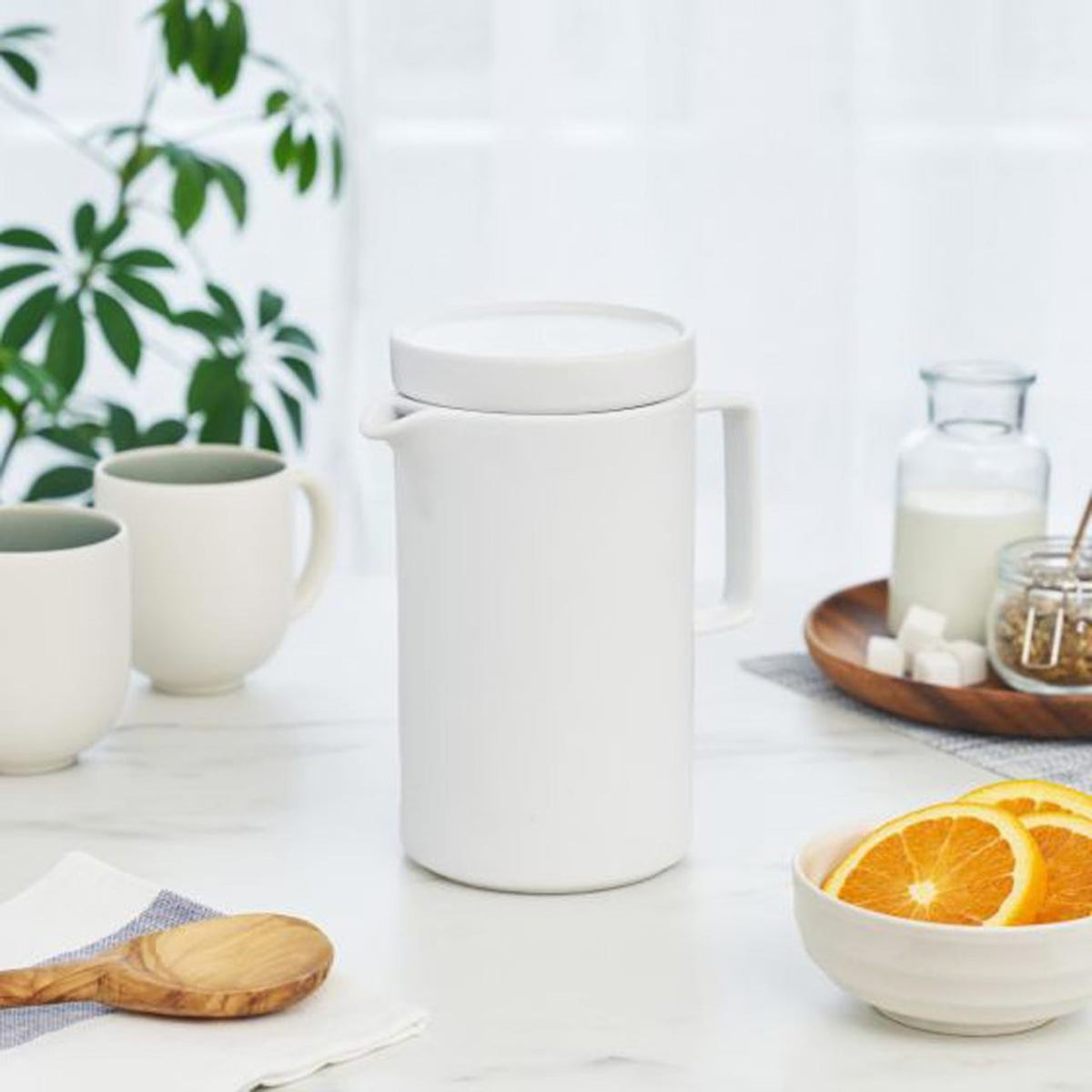 White ceramic tall teapot pitcher on a white surface with mugs, oranges, and milk.