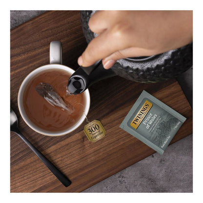 Hand pouring hot tea into a mug of amber colored Prince of Wales Tea on a wood slab with a grey Twining's Prince of Whales teabag package on the table.