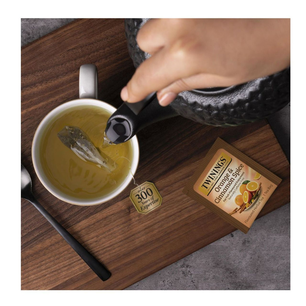 Tea being poured into a cup with a Twinings Orange & Cinnamon Spice tea box on a wooden surface.