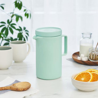 Mint green thermos style teapot on a table with mugs and a bowl of oranges.