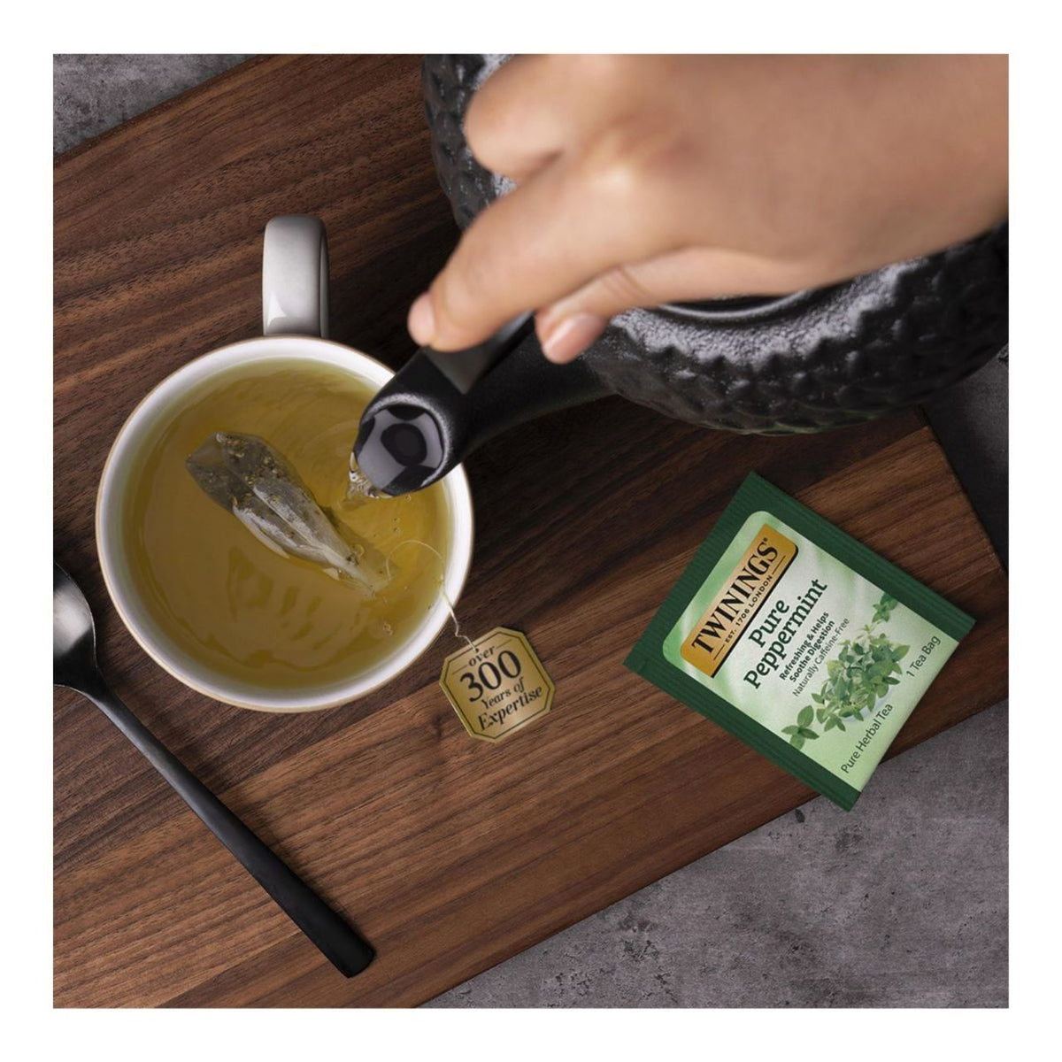 Hand pouring hot tea from black metal teapot into a white cup on a wood panel and a package label for one tea bag of Twinings Pure Peppermint tea