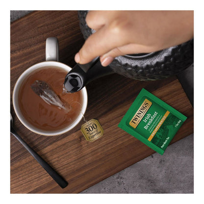 Tea bag being added to a cup of tea with a Twining's tea bag and teapot in the background.