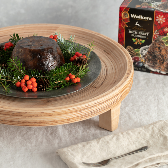 Christmas pudding on a wooden stand with festive decorations and Walkers shortbread packaging in the background.