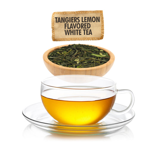 Clear glass cup of tea with a wooden bowl of tea leaves and a tea bag labeled 'Tangiers Lemon Flavored White Tea' on a white background.