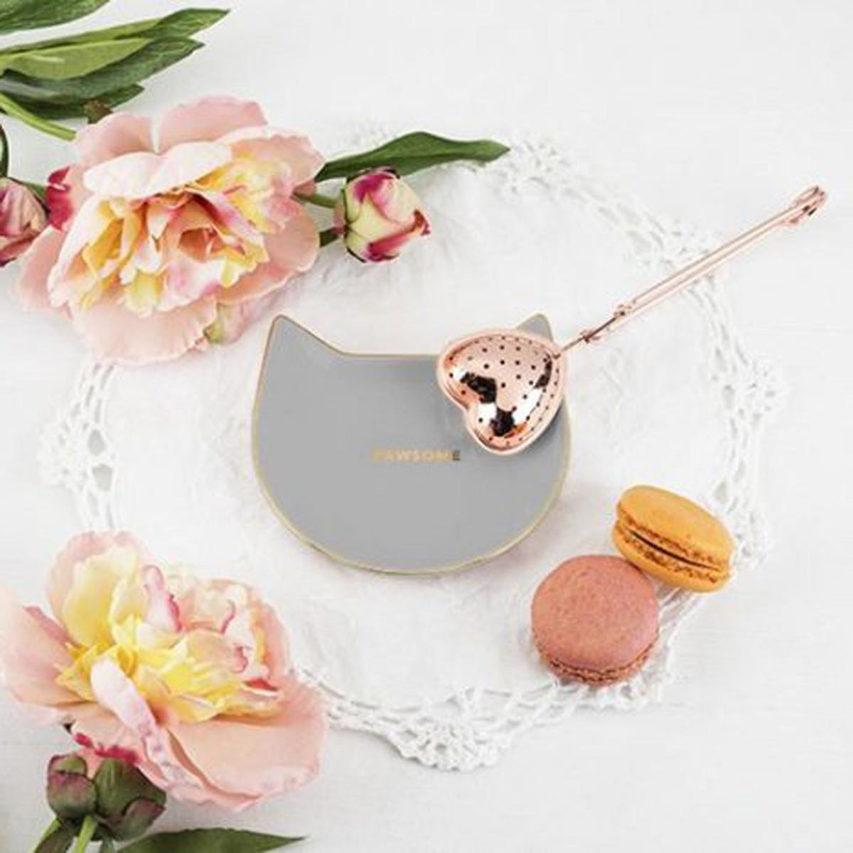 Cat-shaped tea infuser caddy with rose gold accents on a white lace doily with flowers and macarons and a heart shaped tea infuser.
