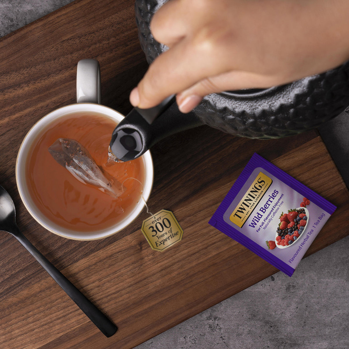 Tea being poured into a cup with a teabag, next to a Twinings Wild Berries tea bag on a wooden surface.