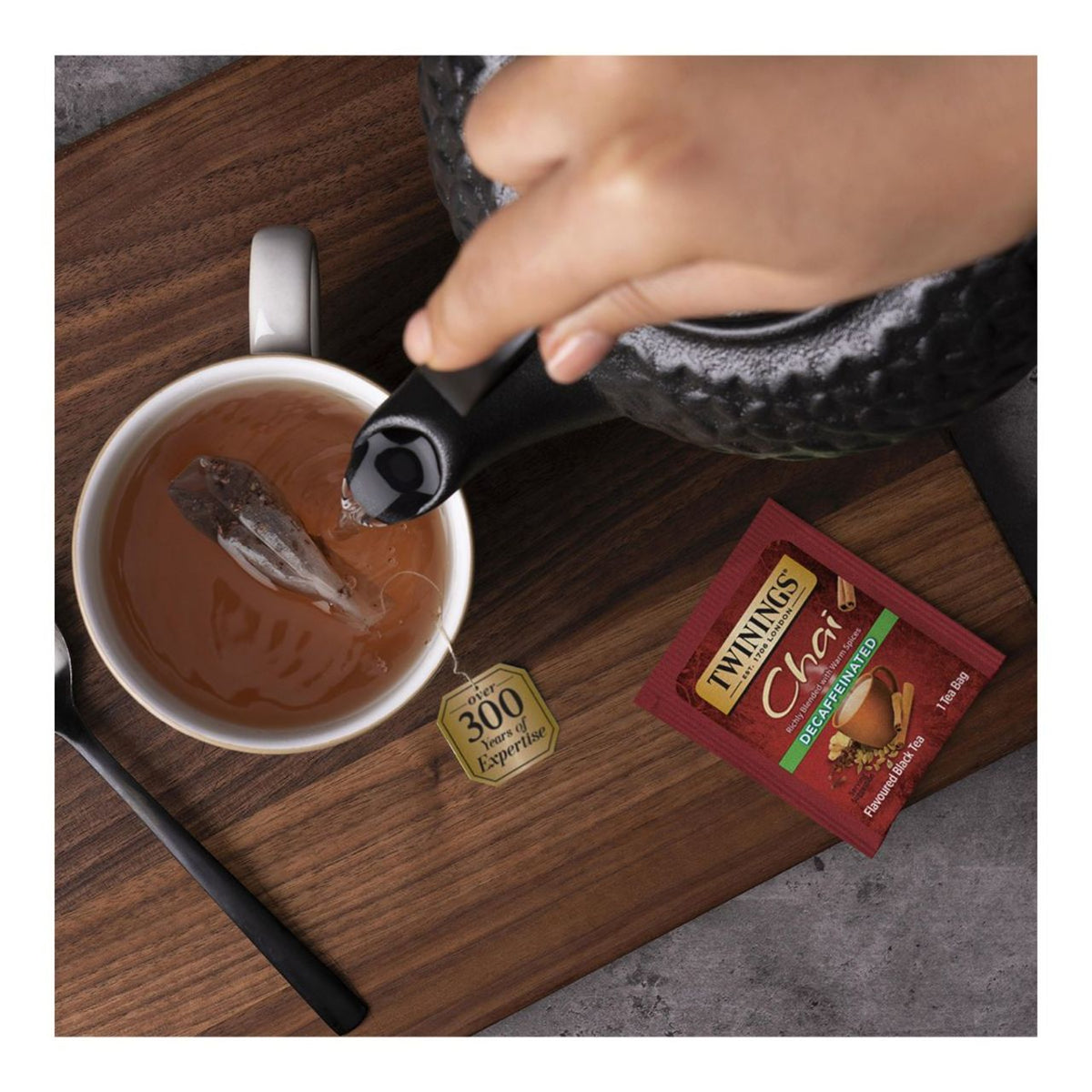Person pouring tea into a cup with a Twining's Chai packet on a wooden surface