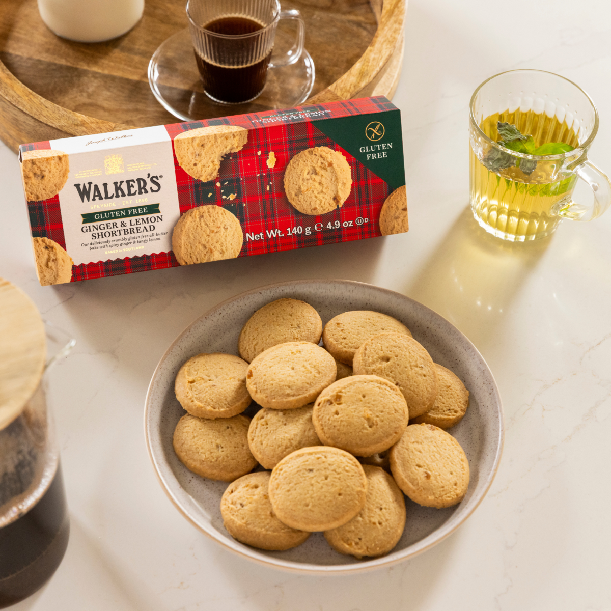 Box of Walker's ginger and lemon shortbread cookies on a table with a bowl of cookies and drinks.