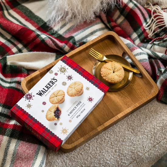 Image of a box of Walker's Mince Pies on a wood tray with a minced pie on a gold plate with fork.  Tray is on a couch cushion with plaid blanket.