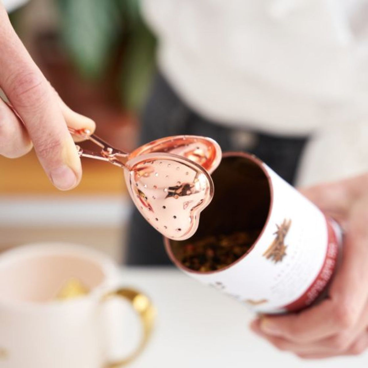 Person using a rose gold heart-shaped scoop infuser to steam tea from a can in a mug.