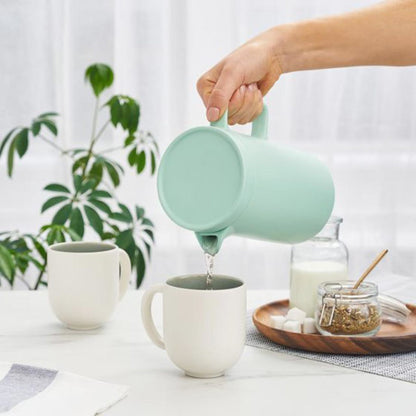 Person pouring tea from a mint green teapot into a white mug on a table with a plant in the background.