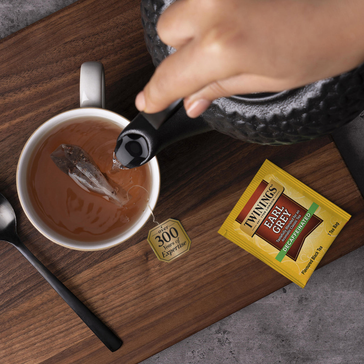 Person pouring tea from a black teapot into a white cup on a wooden surface, with a Thymes Earl Grey tea bag and packaging visible.